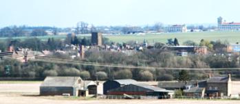 Towcester from Greens Norton showing how the church tower dominates the town. Note the green spire of the Town Hall to the right, Towcester race course in the background, and Lodge Farm in the foreground.